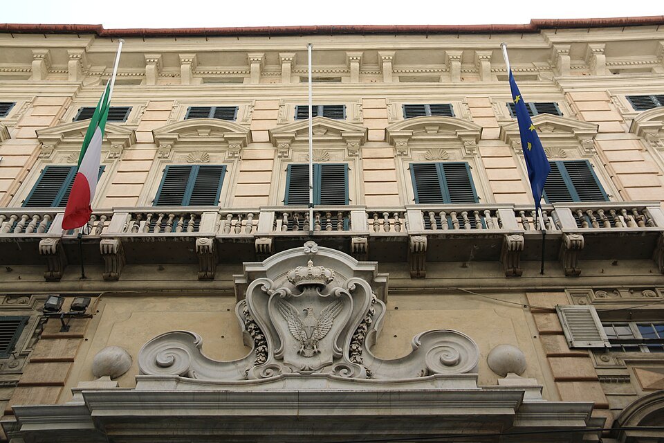 A carved crest over the balcony of a palace front, flags hung at the windows